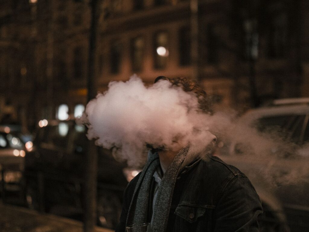 A man exhales a thick cloud of vapor on a city street at night, creating an artistic and dramatic effect.